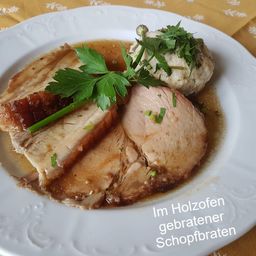 Ein weißer Teller mit geschnittenem Schweinebraten, garniert mit Petersilie, serviert mit Kartoffelpüree, alles in einer Holzschüssel.