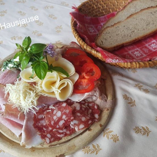 Ein Holzteller mit verschiedenen Fleischsorten, Käse und Tomaten. Ein Korb mit Brot und einem roten Tuch befindet sich nebenan.