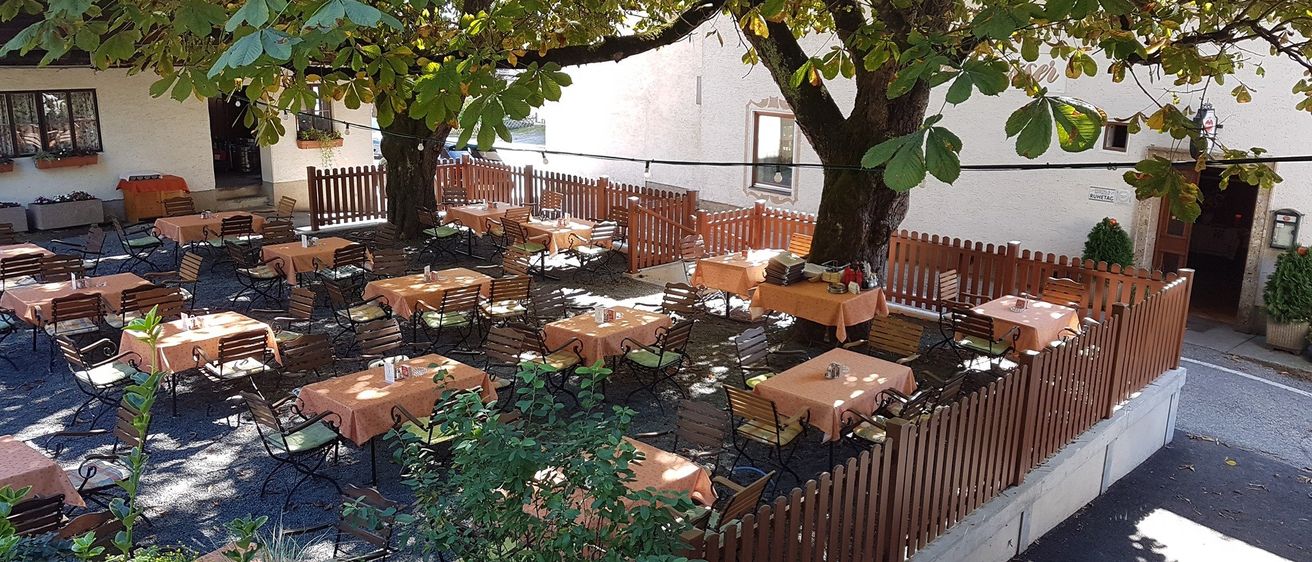 Ein Outdoor-Café mit Tischen und Stühlen unter großen Bäumen, umgeben von Holzzäunen. Im Hintergrund steht ein Gebäude.