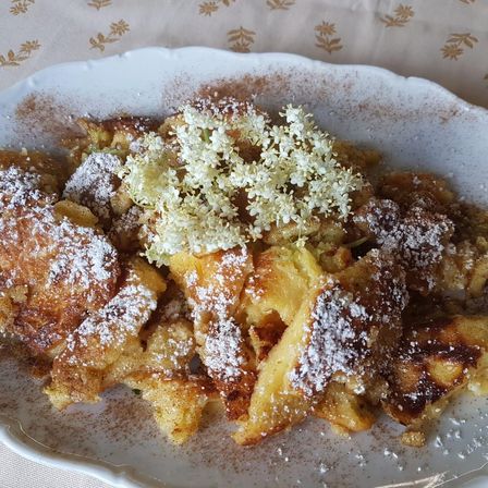 Ein Teller mit gebratenen Kartoffelpuffern, bestreut mit Puderzucker und kleinen weißen Blumen darauf. Das Gericht ist mit Zimt bestreut.
