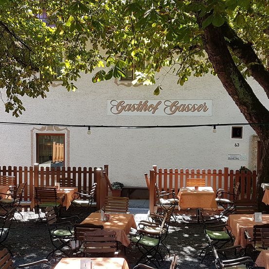 Terrassenbereich des Gasthofs Gasser, einem Restaurant mit Tischen und Stühlen unter einem Baum. An der Wand befindet sich ein Schild mit dem Namen Gasthof Gasser.