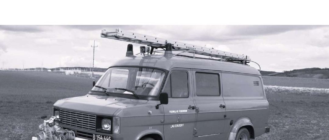 Ein Schwarz-Weiß-Foto eines Ford Transit-Vans mit einer Leiter oben, der auf einem Grasfeld geparkt ist.