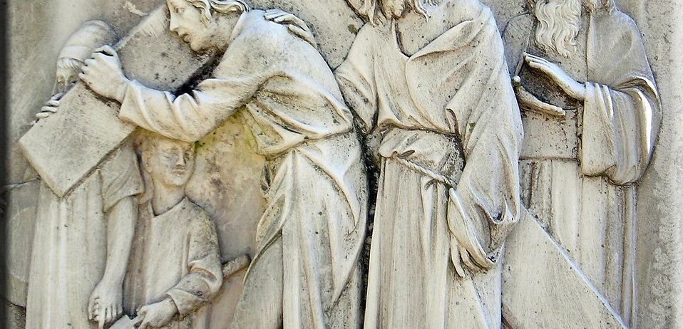 Ein Steinrelief zeigt Jesus, der das Kreuz trägt, unterstützt von einem Mann, während zwei Figuren beobachten. Die Szene fängt den Moment von Christi Weg zum Kalvarienberg ein.