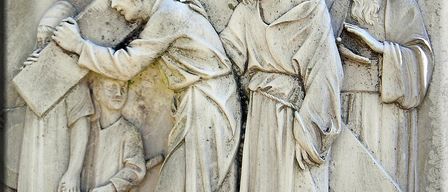 Ein Steinrelief zeigt Jesus, der das Kreuz trägt, unterstützt von einem Mann, während zwei Figuren beobachten. Die Szene fängt den Moment von Christi Weg zum Kalvarienberg ein.
