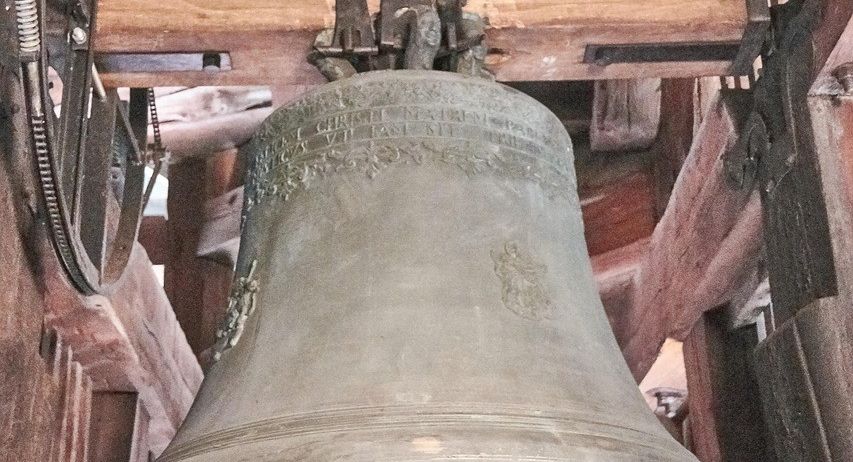 Eine große Glocke hängt von Holzbalken in einem Gebäude. Die Glocke hat aufwendige Verzierungen und einen Metallschlägel.