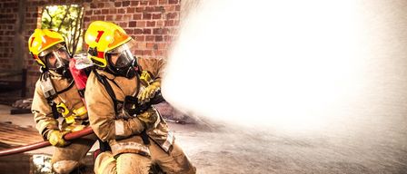 A firefighter, wearing a yellow helmet, is kneeling and spraying water from a hose in a brick building.