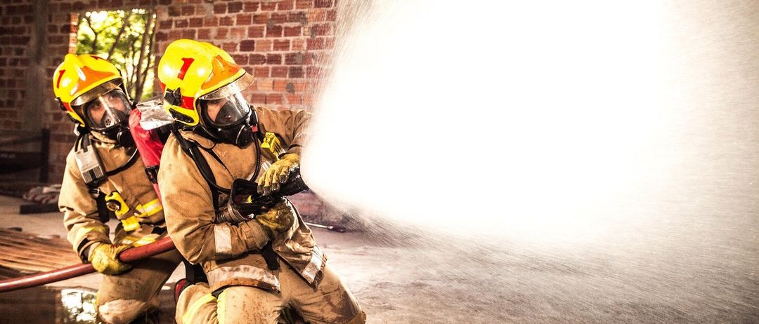 Ein Feuerwehrmann, der einen gelben Helm trägt, kniet und sprüht Wasser aus einem Schlauch in einem Backsteingebäude.