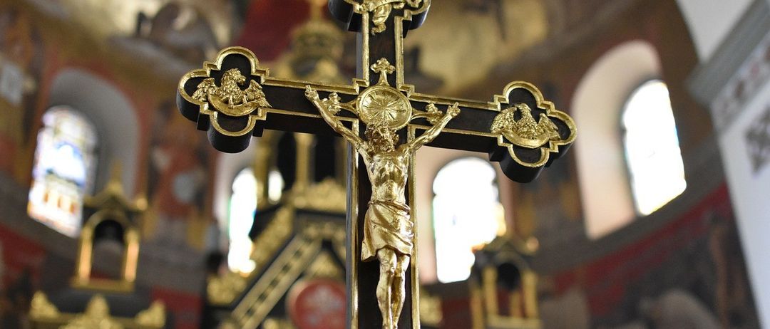 Ein goldenes Kruzifix mit Jesus am Kreuz steht in einer Kirche, mit kunstvollen Verzierungen am Kreuz und goldenen Akzenten. Dahinter zeigt der Kircheninnenraum gewölbte Fenster und kunstvolle Dekorationen.