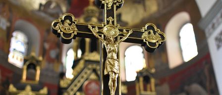 Ein goldenes Kruzifix mit Jesus am Kreuz steht in einer Kirche, mit kunstvollen Verzierungen am Kreuz und goldenen Akzenten. Dahinter zeigt der Kircheninnenraum gewölbte Fenster und kunstvolle Dekorationen.