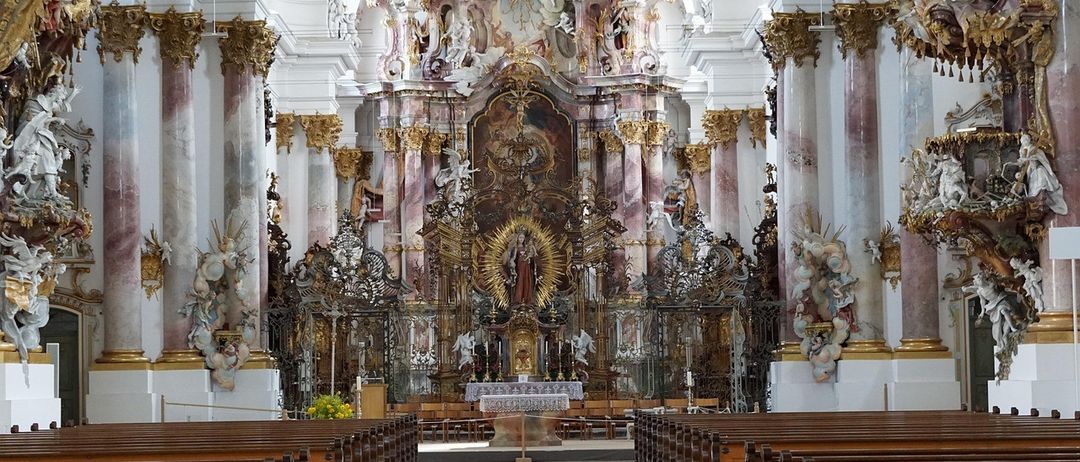 Ein großer, reich verzierter Altar steht in der Mitte einer Kirche mit Statuen und kunstvollen Schnitzereien, umgeben von goldenen Säulen. Der Altar hat eine zentrale Statue und eine goldene Sonnenblume. Die Kirche hat eine hohe Decke mit detaillierten Fresken.