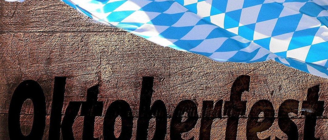 Das Bild zeigt ein blau-weiß kariertes Tuch auf einer Holzoberfläche mit dem Wort Oktoberfest in fetten schwarzen Buchstaben geschrieben.