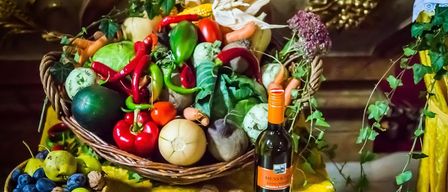 A wicker basket filled with a colorful assortment of vegetables, including peppers, tomatoes, corn, and carrots, sits on a yellow tablecloth. Next to it, a bottle of wine and several walnuts are also visible.