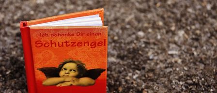 A small red book titled 'I give you a guardian angel' lies on the ground. The cover features a small angel with wings and a serious expression.