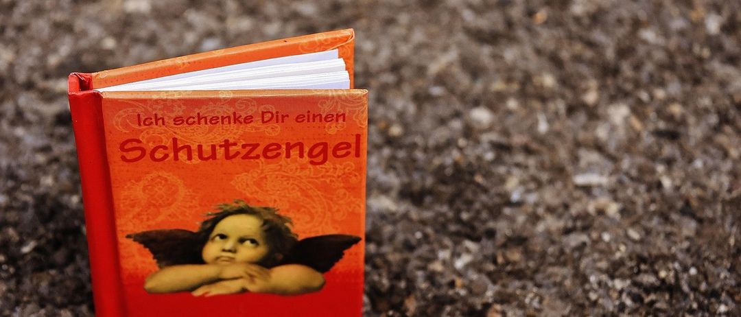Ein kleines rotes Buch mit dem Titel 'Ich schenke Dir einen Schutzengel' liegt auf dem Boden. Das Cover zeigt einen kleinen Engel mit Flügeln und einem ernsten Ausdruck.