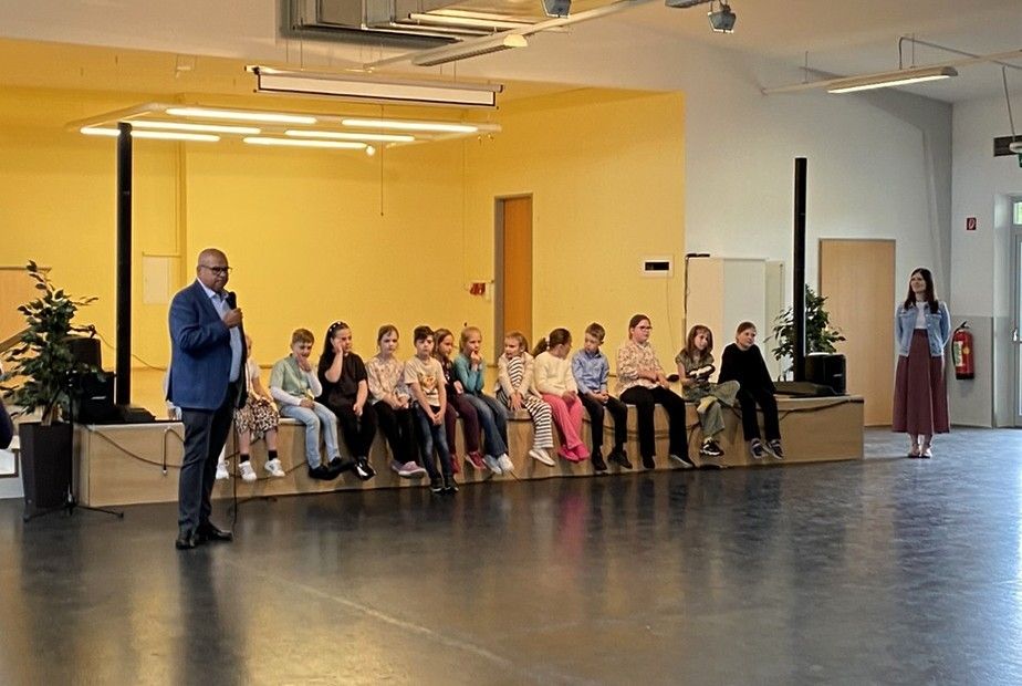 Ein Mann in einem blauen Anzug steht mit einem Mikrofon in einer Halle. Kinder sitzen auf einer Bank vor ihm.