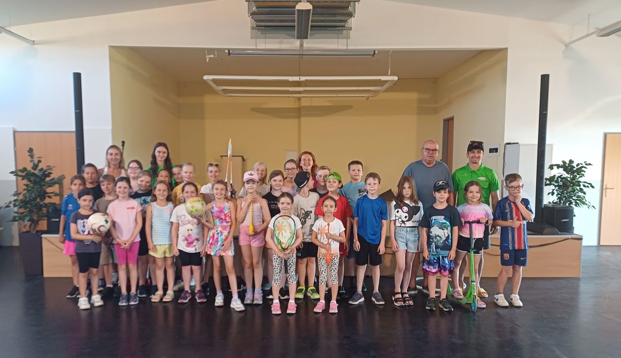 Eine Gruppe von Kindern und Erwachsenen in einem Saal, alle halten Tennisschläger, mit zwei Männern, die davor stehen. Die Kinder tragen verschiedene Outfits und die beiden Männer tragen Brillen. Der Saal hat gelbe Wände und einen schwarzen Boden.