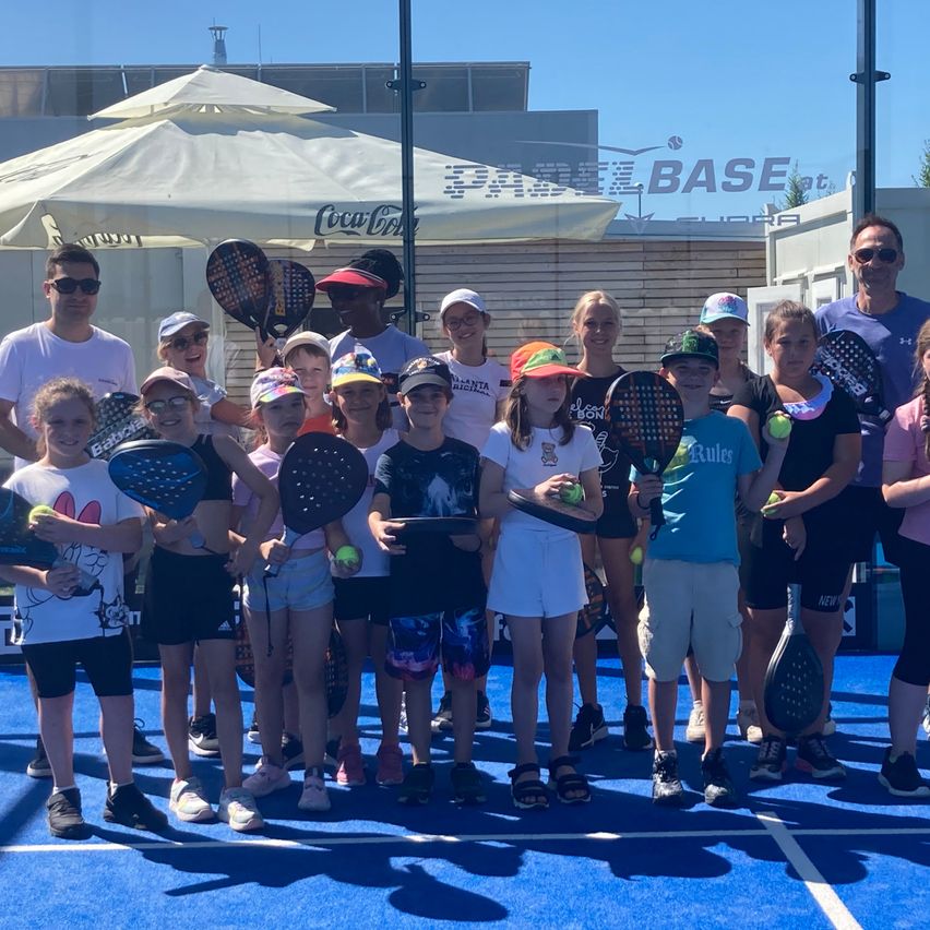 Eine Gruppe von Kindern und Erwachsenen, die Tennisschläger und Tennisbälle halten, posiert für ein Foto auf einem blauen Platz unter einem Baldachin.
