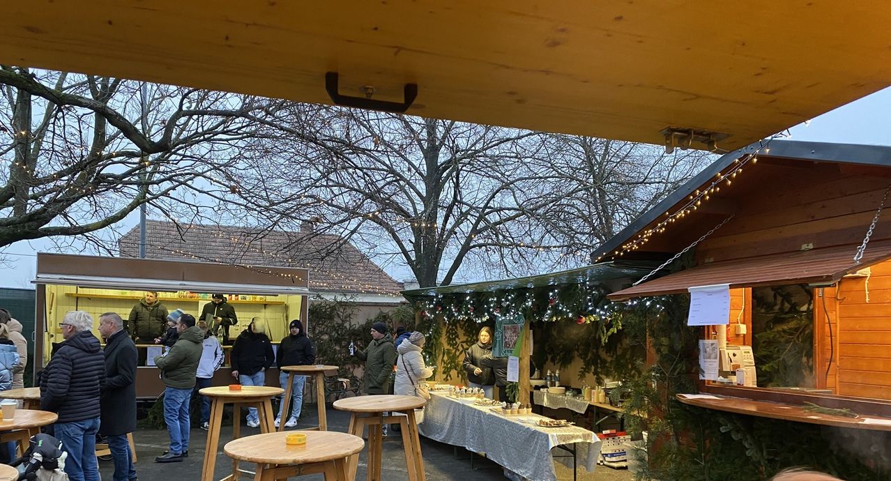 Eine Gruppe von Menschen steht unter einem Vordach mit einer Weihnachtsdekoration. Sie betrachten das Essen auf dem Tisch. Der Tisch hat ein weißes Tuch und einige Teller, Schüsseln und Flaschen. Vor dem Tisch stehen zwei Hocker. Es gibt einen blattlosen Baum und ein Gebäude mit einem Dach.