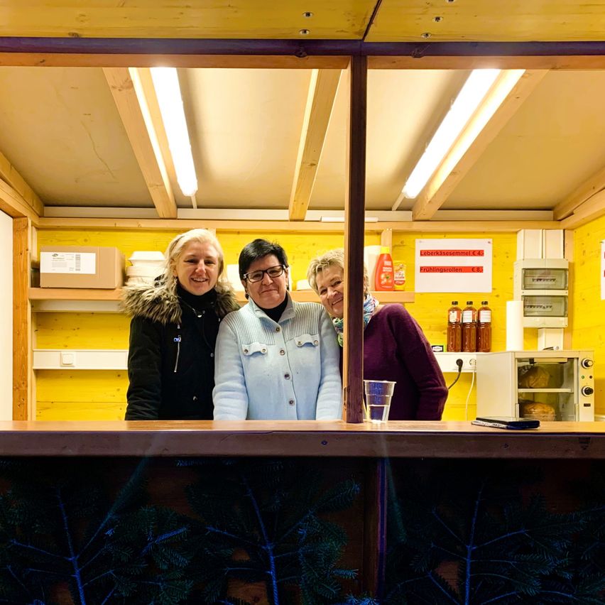 Drei Frauen stehen in einem kleinen, hell beleuchteten Raum und lächeln. Ein Glas Wasser steht auf einer Holztheke, und Regale mit Gegenständen sind im Hintergrund zu sehen.