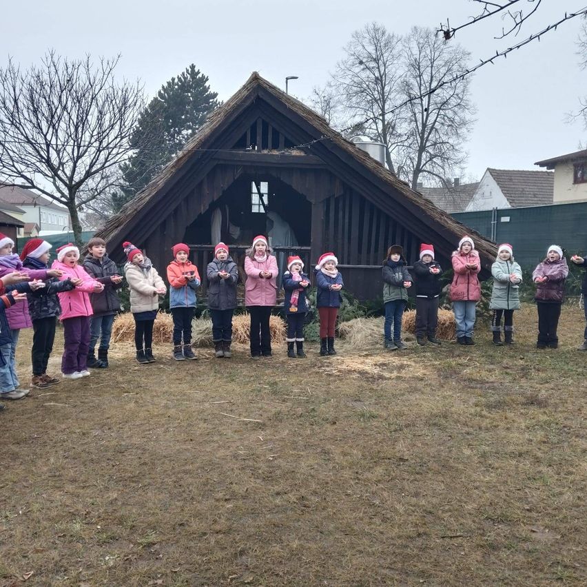 Eine Gruppe von Kindern in Winterkleidung steht in einem Kreis vor einem kleinen Holzhaus mit Strohdach.