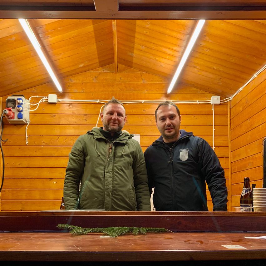 Zwei Männer stehen in einer Holzstruktur und lächeln für ein Foto. Hinter ihnen sind Holzwände und eine Decke mit Beleuchtung zu sehen.