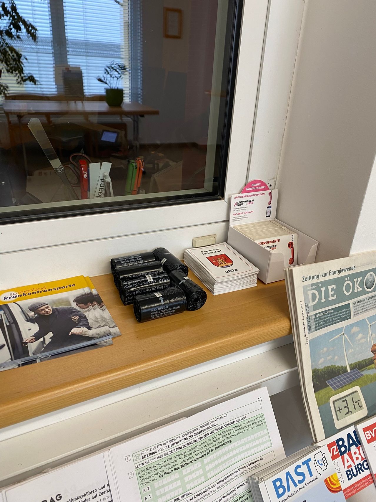 Ein Holzregal hält Broschüren, Batterien und Karten. Eine Zeitung mit einem Mann und Windrädern ist ebenfalls vorhanden. Durch das Fenster ist ein Schreibtisch mit einer Topfpflanze zu sehen.