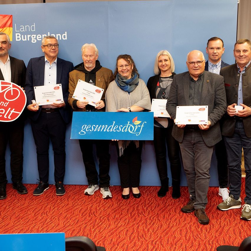 Eine Gruppe von Menschen steht gemeinsam vor einem Banner mit der Aufschrift 'Gesundes Dorf'. Sie halten Urkunden in den Händen und einige haben Fahrräder. Ein blauer Wand mit dem Text 'Land Burgenland' ist hinter ihnen.