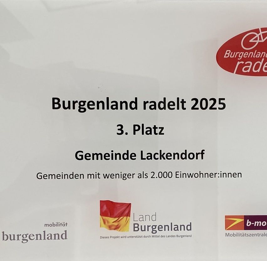 Ein weißes Papier zeigt ein rotes Logo mit dem Text 'Burgenland radelt 2025' und darunter '3. Platz Gemeinde Lackendorf Gemeinden mit weniger als 2.000 Einwohner:innen'. Eine rote Flagge mit einem gelben und schwarzen Design ist auf der linken Seite. Ein blaues Logo mit dem Text 'b-mobilitatszentrum' ist auf der rechten Seite.