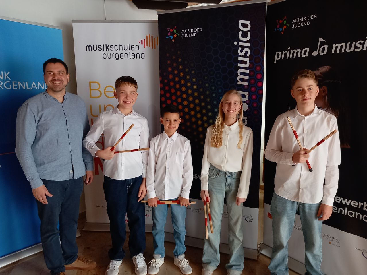 Four children and an adult man are posing for a photo in a room, holding drumsticks. Behind them, there are banners with music-related logos and text.
