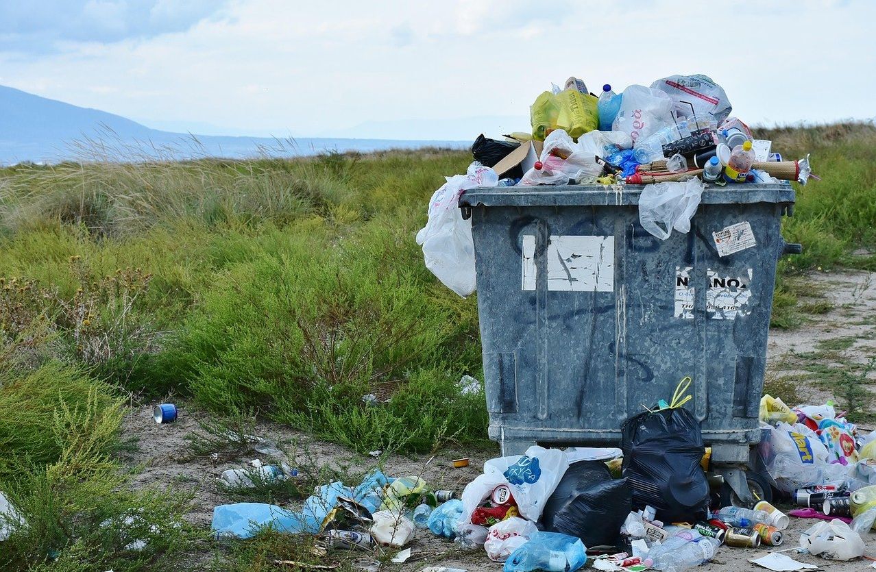 A large trash container filled with plastic waste, including bottles, bags, and other garbage, is overflowing on a grassy field near a body of water.