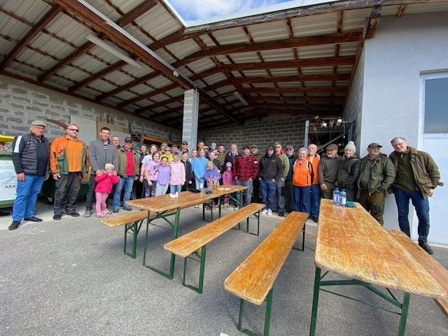 A group of people, including adults and children, are standing and sitting around wooden benches under a metal roof. Some of them are wearing hats and sunglasses. The area is surrounded by a white wall.