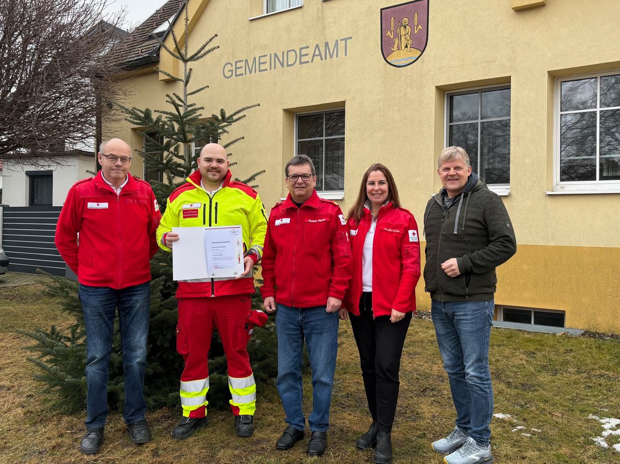 Fünf Personen stehen vor einem Gebäude mit dem Schild 'GEMEINDEAMT'. Einer hält ein Zertifikat. Das Gebäude hat zwei Fenster.