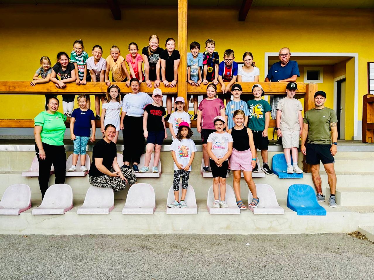 Eine Gruppe von Kindern und Erwachsenen posiert für ein Foto in einem Stadion, wobei einige auf den Tribünen stehen und andere sich an den Geländern lehnen.