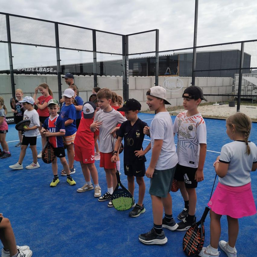 Eine Gruppe von Kindern in Sportkleidung steht in einer Reihe auf einem blauen Tennisplatz und hält Tennisschläger in der Hand.