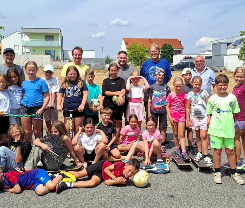 Eine Gruppe von Kindern und Erwachsenen posiert für ein Foto in einem Park. Einige halten Sportgeräte wie Fußballbälle und Skateboards. Im Hintergrund sind Häuser und Bäume zu sehen.