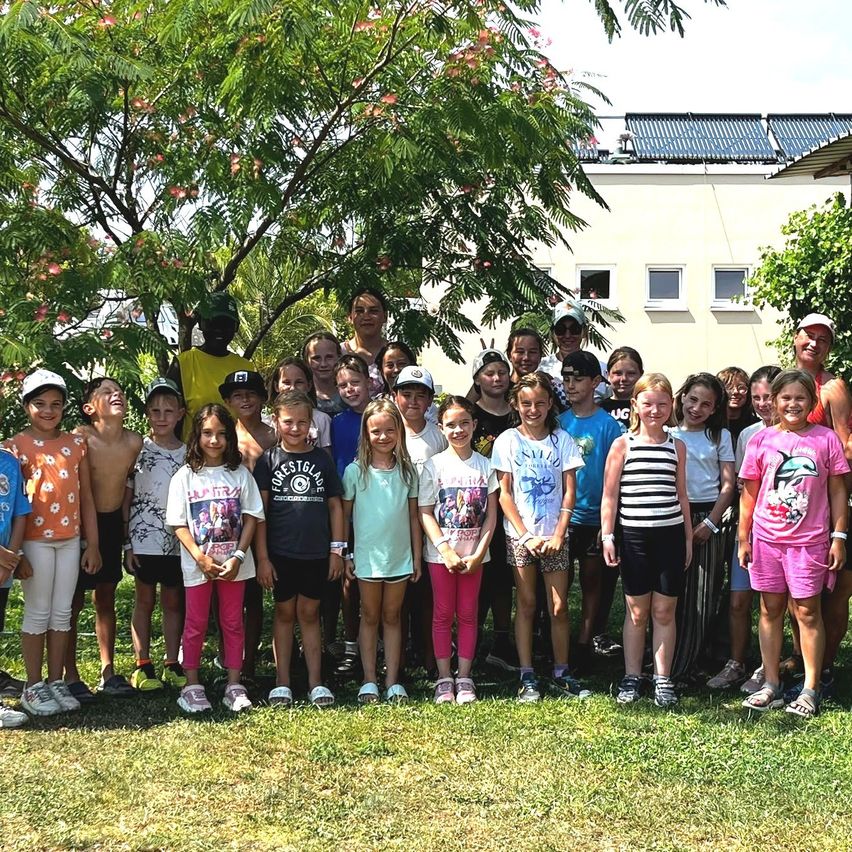 Eine Gruppe von Kindern und Erwachsenen steht auf einer Rasenfläche, mit einem Gebäude mit Solarpanels im Hintergrund.