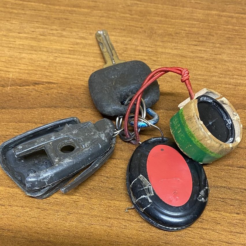Drei Autoschlüssel liegen auf einer Holzoberfläche. Ein Schlüssel ist beschädigt und hat einen roten Kreis, ein anderer ist in braune Papier gewickelt.