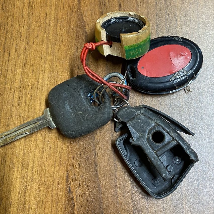 Ein Schlüsselsatz mit einem schwarzen Plastikschlüssel, einem rot bemalten Knopf und einem hölzernen und grünen Objekt auf einer Holzoberfläche.