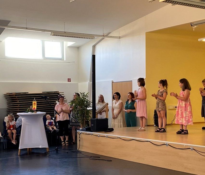 A group of young girls stand on stage, possibly for a ceremony. A woman and a child sit on the left. Others stand behind the stage. A potted plant is on the stage.