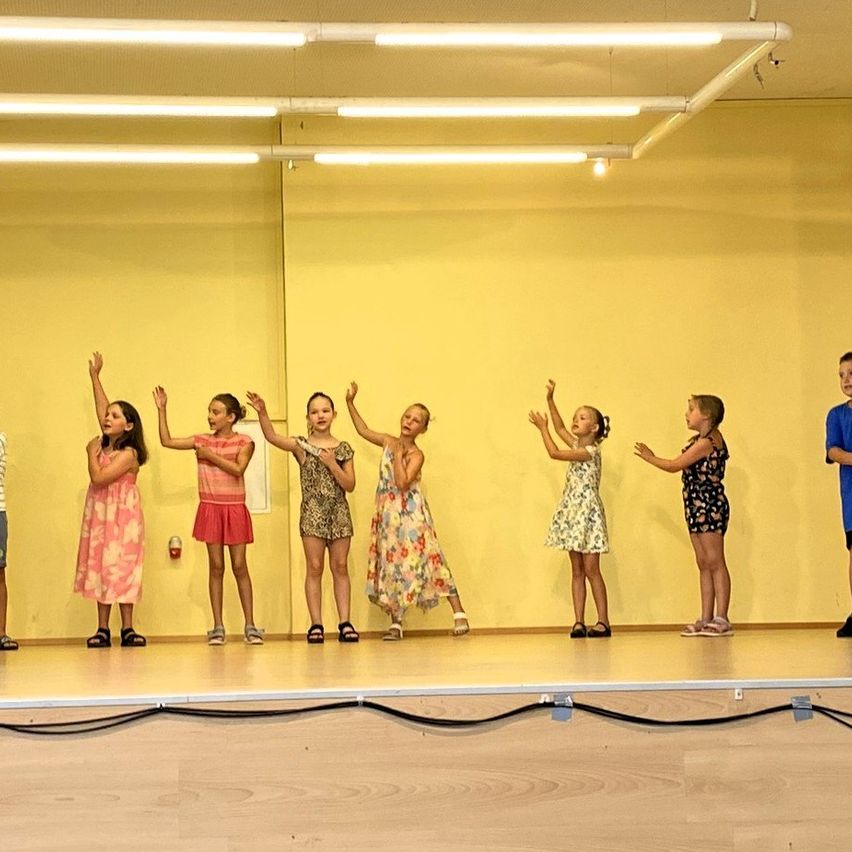 A group of young girls perform a coordinated dance routine on a stage with yellow walls and a mirror. They wear various dresses and have their hands raised.
