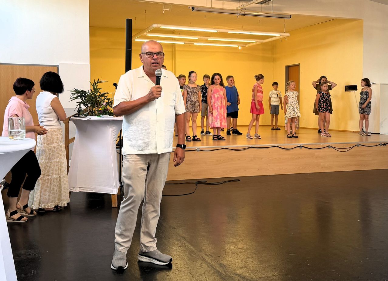 A man stands on stage with a microphone, addressing a group of children lined up in a room. A plant and a table are nearby.