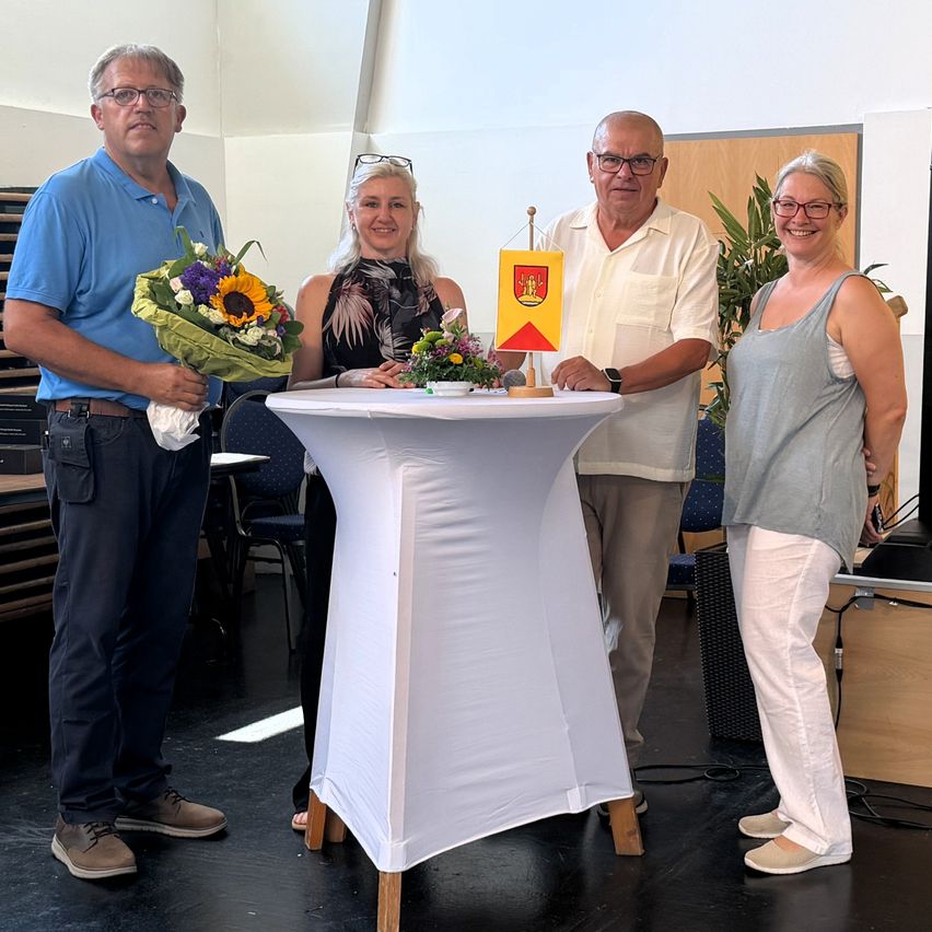 Four people stand around a table with flowers and a flag. The man holds a bouquet, and the woman wears glasses. They smile for a photo.