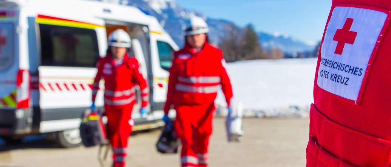 Zwei Personen in roten Uniformen, möglicherweise Rettungskräfte, gehen auf einer verschneiten Straße mit Bergen im Hintergrund.