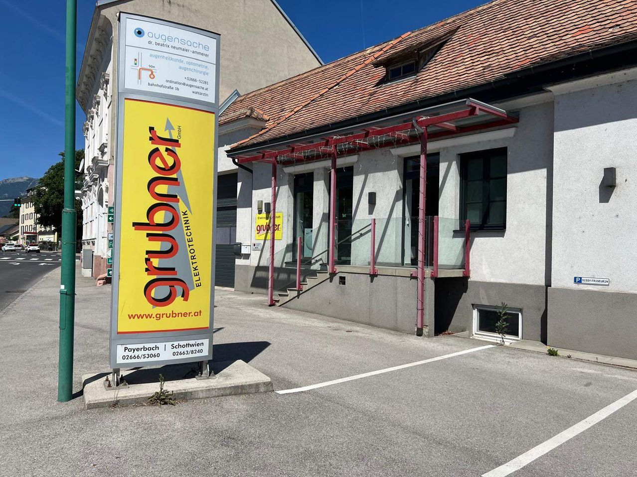 Ein gelbes Schild für Grubner steht vor einem Gebäude mit einem Ziegeldach, Glastüren und einer Treppe. Das Schild enthält eine Website und Kontaktinformationen.
