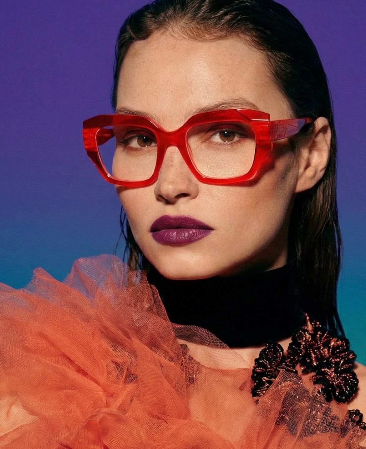 Eine Frau mit roten Katzenaugenbrille und dunklem Lippenstift. Sie hat dunkles Haar und trägt eine schwarze Halskette und ein orangefarbenes Rüschenoberteil.