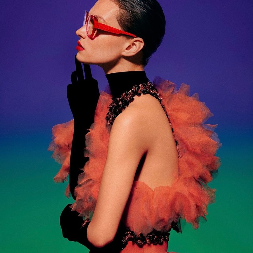 Eine Frau in einem roten Kleid mit Rüschen und schwarzen Handschuhen posiert mit einem roten Rahmen im Gesicht vor einem lila-grünen Hintergrund.