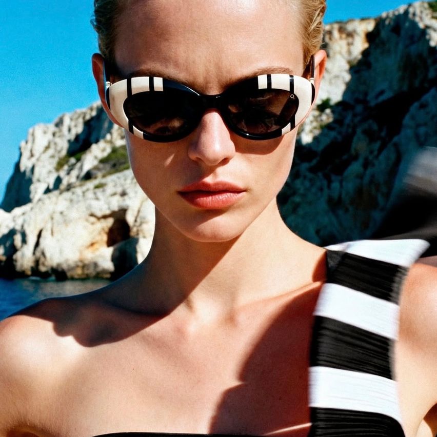 A woman wearing sunglasses with a black and white striped pattern stands outside. She is looking forward with her lips slightly open. Behind her, there is a large rock formation.