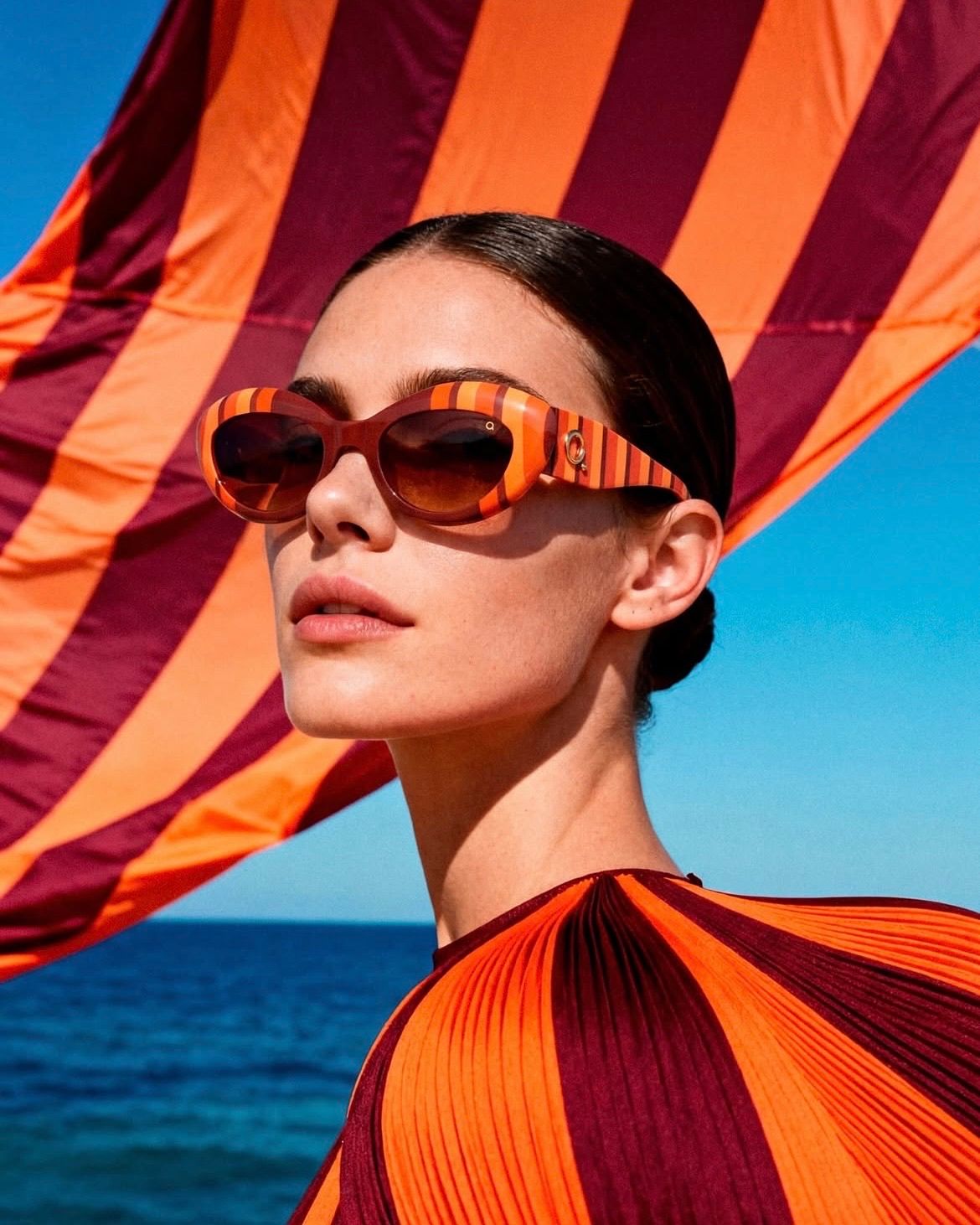 A woman stands by the ocean, wearing sunglasses with an orange and maroon striped pattern. Her hair is tied back. The sky is blue, and she is looking towards the camera.