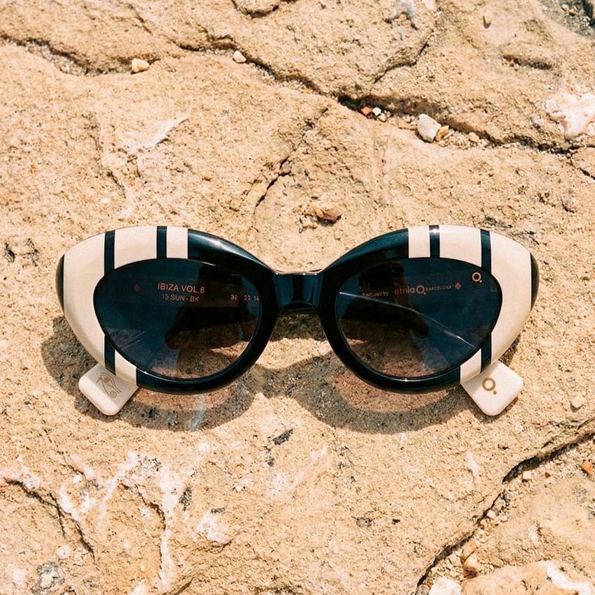 A pair of black and white cat-eye sunglasses with Ibiza Vol.6 branding lie on sandy ground.