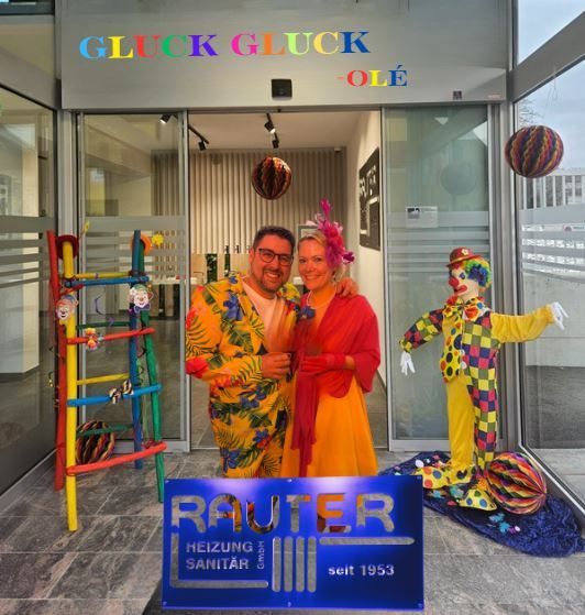 Ein Mann und eine Frau stehen vor einem Geschäft mit bunten Dekorationen und Luftballons. Das Ladenschild steht in leuchtenden Farben für Gluck Gluck Ole. Sie sind in leuchtenden Clown-Kostümen gekleidet. Der Laden scheint Rauter zu sein, das seit 1953 in Betrieb ist.